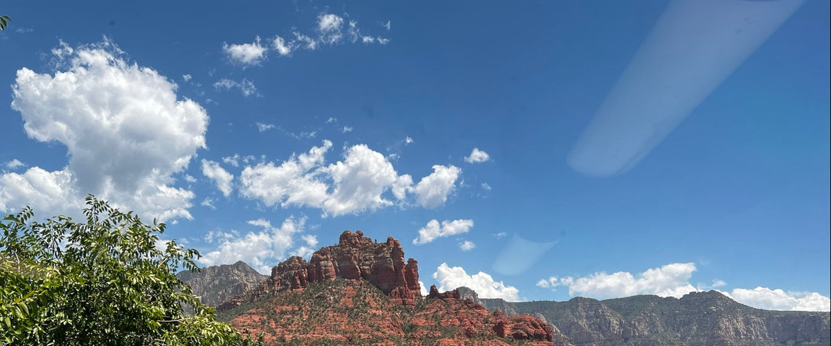 A scenic landscape photograph of Sedona, Arizona, featuring a prominent red rock butte rising above a lush green forest under a bright blue sky with white clouds.